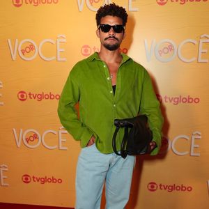 Lucas Leto aposta em um estilo casual colorido com camisa de veludo cotelê verde-vibrante usada levemente aberta sobre regata preta, combinada com calça jeans clara de modelagem reta. O look traz óculos de sol pretos clássicos, bolsa tiracolo preta e mocassins pretos com fivela