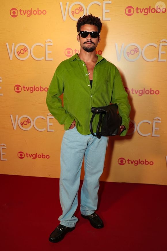 Lucas Leto aposta em um estilo casual colorido com camisa de veludo cotelê verde-vibrante usada levemente aberta sobre regata preta, combinada com calça jeans clara de modelagem reta. O look traz óculos de sol pretos clássicos, bolsa tiracolo preta e mocassins pretos com fivela