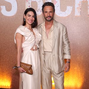 Mel Fronckowiak veste um modelo midi acetinado creme com franzidos volumosos no decote, recortes laterais na cintura e fenda discreta, combinado a uma clutch de couro caramelo. Ao seu lado, Rodrigo Santoro usa alfaiataria bege com textura sutil de riscas, camisa de gola aberta no mesmo tom e mocassins em camurça marrom