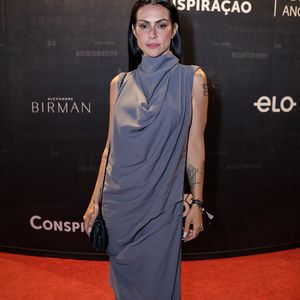 Cleo trouxe uma leitura contemporânea e sofisticada com vestido longo em tom cinza, de modelagem fluida e assimétrica, com gola alta drapeada e recortes que acompanham o corpo, evidenciando suas tatuagens; o styling monocromático, aliado à maquiagem marcante e cabelos alinhados, reforça uma estética moderna e imponente no tapete vermelho