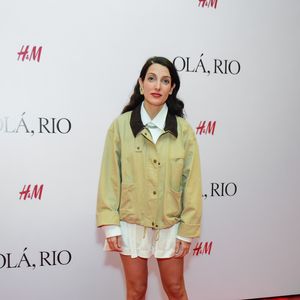 Clarice Falcão montou um look que mistura referências colegiais e utilitárias. A cantora utilizou uma jaqueta estilo bomber em tom cáqui com gola de veludo cotelê marrom, sobreposta a uma camisa branca fechada até o colarinho e uma saia curta branca plissada. A composição foi finalizada com meias brancas aparentes e mocassins pretos de bico arredondado com detalhes dourados