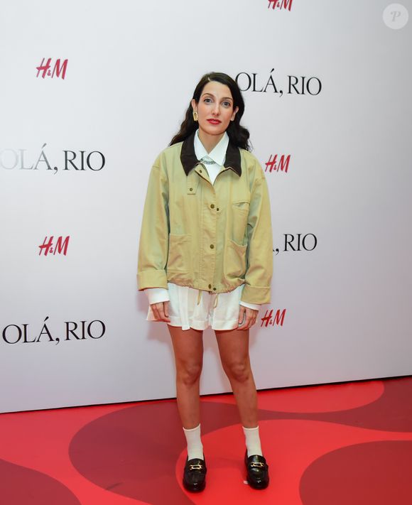 Clarice Falcão montou um look que mistura referências colegiais e utilitárias. A cantora utilizou uma jaqueta estilo bomber em tom cáqui com gola de veludo cotelê marrom, sobreposta a uma camisa branca fechada até o colarinho e uma saia curta branca plissada. A composição foi finalizada com meias brancas aparentes e mocassins pretos de bico arredondado com detalhes dourados