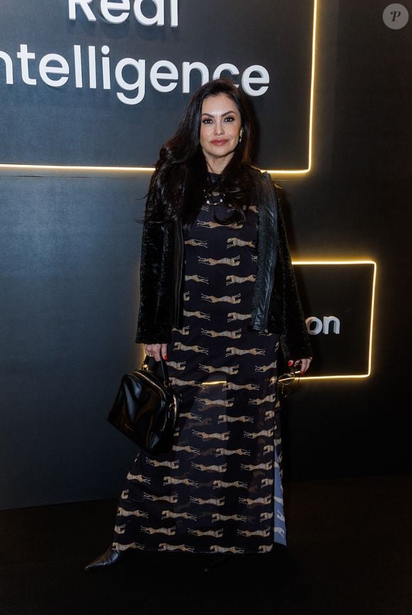 Renata Bráz comparece ao evento com uma combinação de texturas e estampas marcantes. Ela exibe um vestido longo de base escura e tecido fluido, inteiramente decorado com uma padronagem repetida de leopardos em tons dourados e beges. A barra do vestido revela uma leve transparência sob a luz. Para se proteger do frio, Liége sobrepõe a peça com um casaco preto confeccionado em material que imita pele, apresentando debruns frontais com acabamento em couro sintético liso. Como acessórios, ela carrega uma bolsa de mão estruturada em couro preto brilhante e calça botas de bico fino na mesma cor