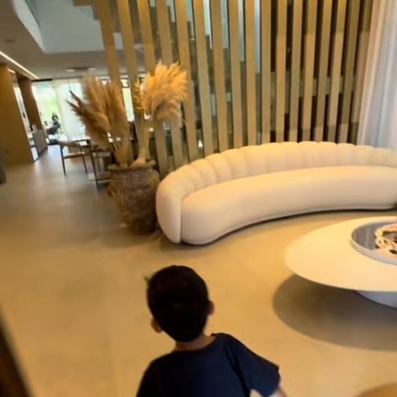 Na sala, um sofá curvo imponente se torna o grande protagonista do décor, dialogando com a mesa de centro orgânica e com o painel ripado vertical que separa os ambientes sem bloquear a luz natural