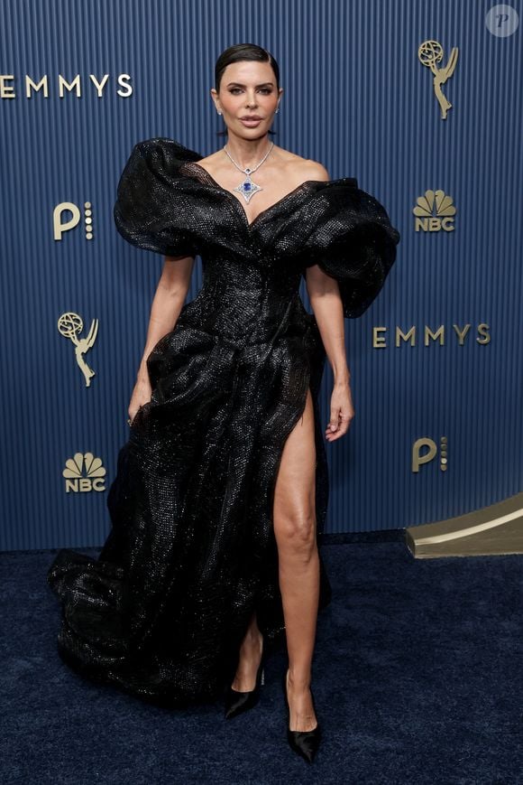 Lisa Rinna aposta em um imponente vestido preto de tecido reluzente, destacado pelo decote ombro a ombro estruturado com mangas curtas altamente volumosas e esculpidas. O modelo com caimento fluido conta com uma fenda profunda que exibe a perna e scarpins pretos clássicos. Um colar com gema azul central completa o visual ousado