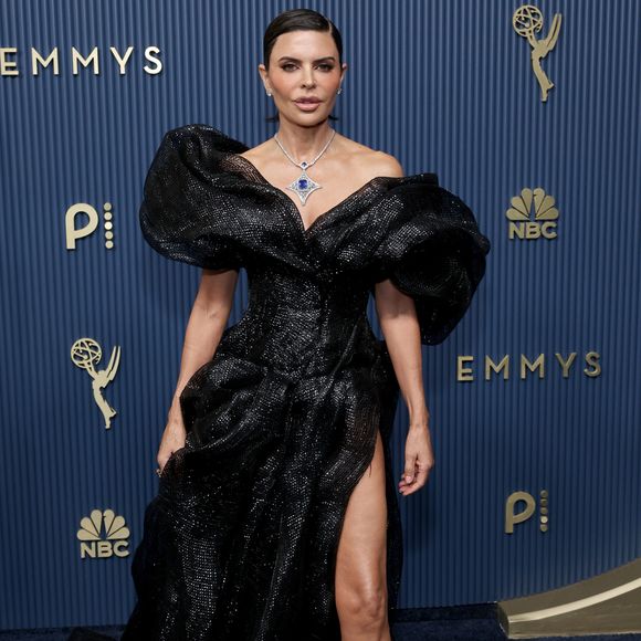 Lisa Rinna aposta em um imponente vestido preto de tecido reluzente, destacado pelo decote ombro a ombro estruturado com mangas curtas altamente volumosas e esculpidas. O modelo com caimento fluido conta com uma fenda profunda que exibe a perna e scarpins pretos clássicos. Um colar com gema azul central completa o visual ousado