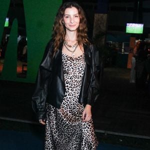 Giselle Batista aposta em um slip dress midi acetinado com estampa de animal print de leopardo, sobreposto por uma jaqueta oversized de couro preto com modelagem despojada. O visual ganha ares urbanos com botas pretas de cano médio e saltos estruturados, além de um mix de colares de correntes sobrepostos