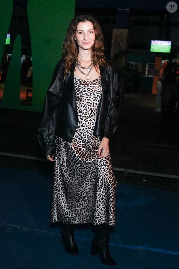 Giselle Batista aposta em um slip dress midi acetinado com estampa de animal print de leopardo, sobreposto por uma jaqueta oversized de couro preto com modelagem despojada. O visual ganha ares urbanos com botas pretas de cano médio e saltos estruturados, além de um mix de colares de correntes sobrepostos