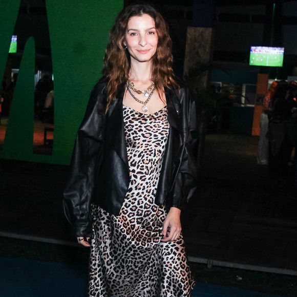 Giselle Batista aposta em um slip dress midi acetinado com estampa de animal print de leopardo, sobreposto por uma jaqueta oversized de couro preto com modelagem despojada. O visual ganha ares urbanos com botas pretas de cano médio e saltos estruturados, além de um mix de colares de correntes sobrepostos