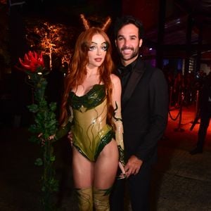 Duda Reis e marido trajam looks temáticos. Duda veste um macacão body de latex verde musgo com detalhes que lembram veias e folhas, botas de cano alto combinando e luvas transparentes com detalhes verdes nas mãos. Ela usa uma maquiagem temática, cabelos ruivos longos com dois pequenos "chifres" de cabelo no topo da cabeça, e segura um bastão ornamentado com folhagem e uma flor vermelha na ponta. O seu marido veste um terno preto clássico com gravata borboleta
