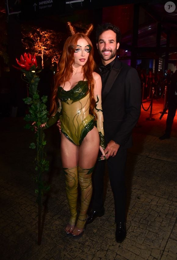 Duda Reis e marido trajam looks temáticos. Duda veste um macacão body de latex verde musgo com detalhes que lembram veias e folhas, botas de cano alto combinando e luvas transparentes com detalhes verdes nas mãos. Ela usa uma maquiagem temática, cabelos ruivos longos com dois pequenos "chifres" de cabelo no topo da cabeça, e segura um bastão ornamentado com folhagem e uma flor vermelha na ponta. O seu marido veste um terno preto clássico com gravata borboleta