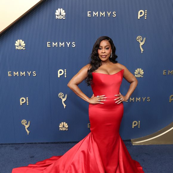 Niecy Nash-Betts veste um modelo sereia vermelho em cetim com corpete tomara que caia de decote arquitetônico estruturado. A peça justa ao corpo se desdobra a partir dos joelhos em uma volumosa cauda circular arrastada pelo tapete. Ela complementa a produção com cabelos ondulados em ondas clássicas e unhas em tom claro