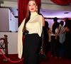 Nicole Bahls entregou uma estética de 'vilã de cinema' clássica, inspirada no glamour das décadas passadas. O visual é centrado em uma blusa off-white de gola alta e caimento assimétrico, com uma das mangas estendida em uma capa fluida. A peça foi combinada a uma saia lápis preta de cintura alta e comprimento midi. Os acessórios foram cruciais para o impacto do look: luvas de ópera em couro preto, uma mini bolsa de corrente, scarpins de verniz com bico extremamente fino e brincos de argola dourados de design robusto. O batom vermelho intenso completou a assinatura visual