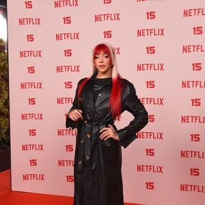 Pabllo Vittar: apresenta um trench coat em pele preta ajustado na cintura com cinto e fivela metálica, sobreposto a uma blusa preta de gola alta, complementado por botas pretas de salto e cabelo longo com franja e pontas em tom vermelho-vibrante