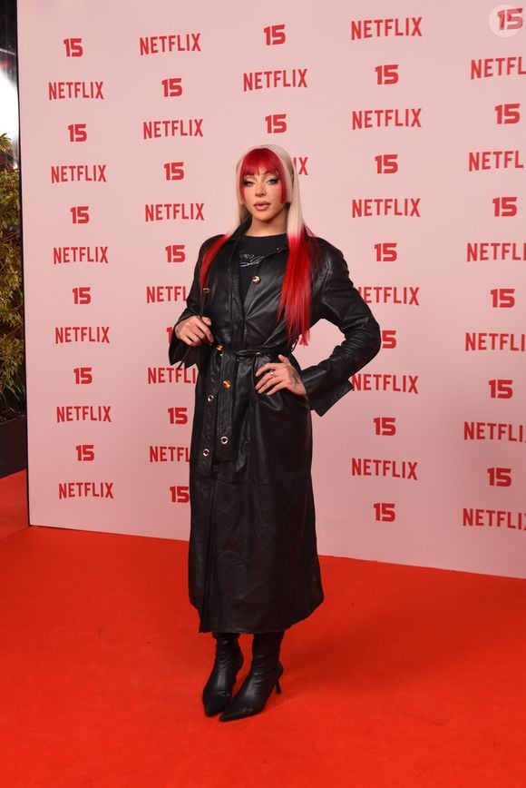 Pabllo Vittar: apresenta um trench coat em pele preta ajustado na cintura com cinto e fivela metálica, sobreposto a uma blusa preta de gola alta, complementado por botas pretas de salto e cabelo longo com franja e pontas em tom vermelho-vibrante