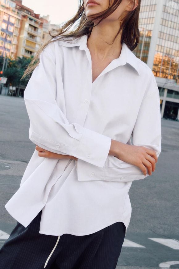 Em branco, a Camisa Básica Oxford da Zara tem fechamento frontal por botões e mangas amplas com acabamento em punho, além de gola de lapela e decote V