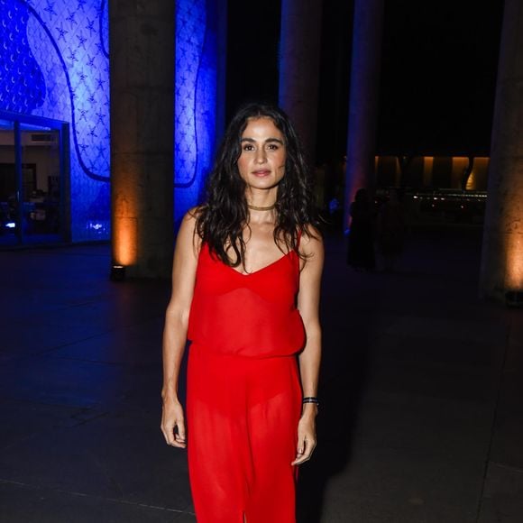 Nanda Costa aposta em um visual de apelo contemporâneo e despretensioso, com vestido vermelho de alças finas e decote em V, confeccionado em tecido leve e levemente translúcido, que cria um efeito fluido ao longo do corpo e ganha movimento com a fenda frontal discreta; a escolha de styling foge do óbvio ao incorporar sapatos 'masculinos' pretos de acabamento polido, quebrando a feminilidade clássica da peça