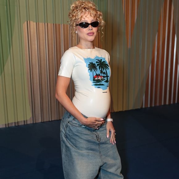 A criadora de conteúdo Malu Borges, conhecida por seus looks experimentais, exibiu sua gravidez com um visual que une o pop ao surrealismo. Ela utilizou uma t-shirt de látex branco da marca Normando, com uma estampa tropical centralizada, ajustada ao corpo para destacar a silhueta da gestação. A parte inferior foi composta por uma saia jeans de modelagem extremamente ampla e desestruturada, criando um jogo de proporções exageradas. Nos pés, as famosas sapatilhas de flores vermelhas tridimensionais adicionaram a textura e o volume que são marcas registradas de seu estilo autêntico