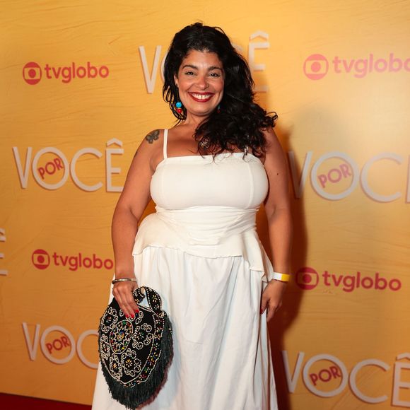 Daniela Fontan aposta em um vestido midi branco monocromático de alças finas, com corpete ajustado e peplum discreto sobre a saia de caimento reto. O destaque fica para a bolsa artesanal com franjas e bordados coloridos, combinada com scarpins de bico fino e salto baixo