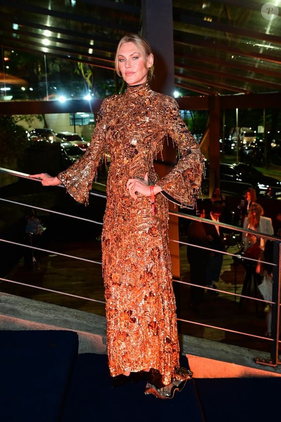Shirley Mallmann: a top model optou por um vestido longo de gola alta e mangas longas estilo boca de sino, inteiramente bordado com paetês, vidrilhos e texturas metálicas em tom bronze/cobre. A produção conta com detalhes de franjas reluzentes e é finalizada com os cabelos presos em um coque alinhado