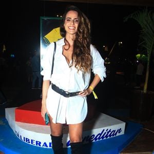 Marcella Fogaça veste uma proposta minimalista e ousada de chemise branca de modelagem ampla usada como minivestido, ajustada na cintura por um cinto largo de couro preto com tachas e fivela metálica. O destaque fashionista fica por conta das botas over-the-knee pretas em camurça, que alongam a silhueta
