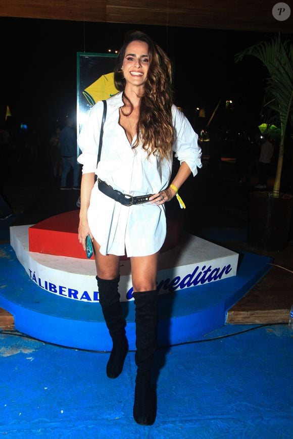 Marcella Fogaça veste uma proposta minimalista e ousada de chemise branca de modelagem ampla usada como minivestido, ajustada na cintura por um cinto largo de couro preto com tachas e fivela metálica. O destaque fashionista fica por conta das botas over-the-knee pretas em camurça, que alongam a silhueta