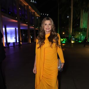 Flávia Alessandra aposta em uma paleta vibrante com um vestido midi em tom mostarda intenso. A peça apresenta drapeados estratégicos ao longo do corpo e um detalhe de tecido alongado que funciona como uma espécie de capa lateral, adicionando fluidez e elegância. A modelagem ajustada valoriza a silhueta, enquanto as sandálias de salto fino e a clutch prateada trazem brilho na medida certa