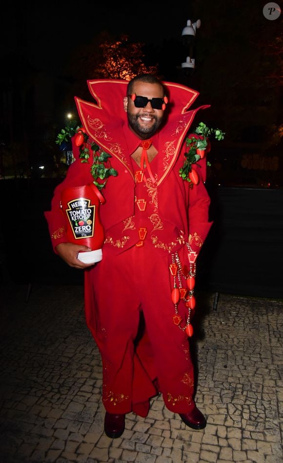 John Drops veste um fato temático encarnado de corte dramático com gola alta estruturada e bordados dourados, inspirado na marca de ketchup Heinz. O fato inclui aplicações de folhagens e tomates de plástico nos ombros e na cintura, óculos de sol pretos com pequenos tomates anexados e um acessório em formato de embalagem gigante de ketchup na mão