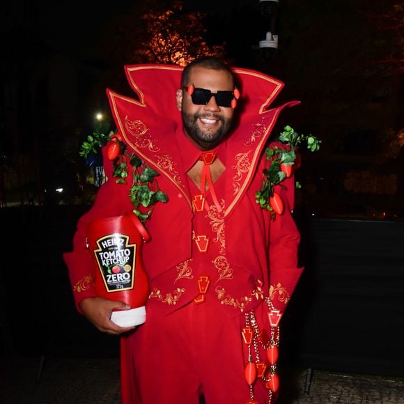 John Drops veste um fato temático encarnado de corte dramático com gola alta estruturada e bordados dourados, inspirado na marca de ketchup Heinz. O fato inclui aplicações de folhagens e tomates de plástico nos ombros e na cintura, óculos de sol pretos com pequenos tomates anexados e um acessório em formato de embalagem gigante de ketchup na mão