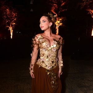 Duda Rubert veste um vestido longo de gala com temática de fada e natureza. A peça possui corpete estruturado tomara que caia e mangas longas em tule transparente, decorados com bordados dourados e aplicações de folhas metálicas e pequenas flores rosa. A saia é fluida e escura, em tom marrom com caimento dramático e arranjo de folhas na cintura, harmonizando com o arranjo folheado no cabelo