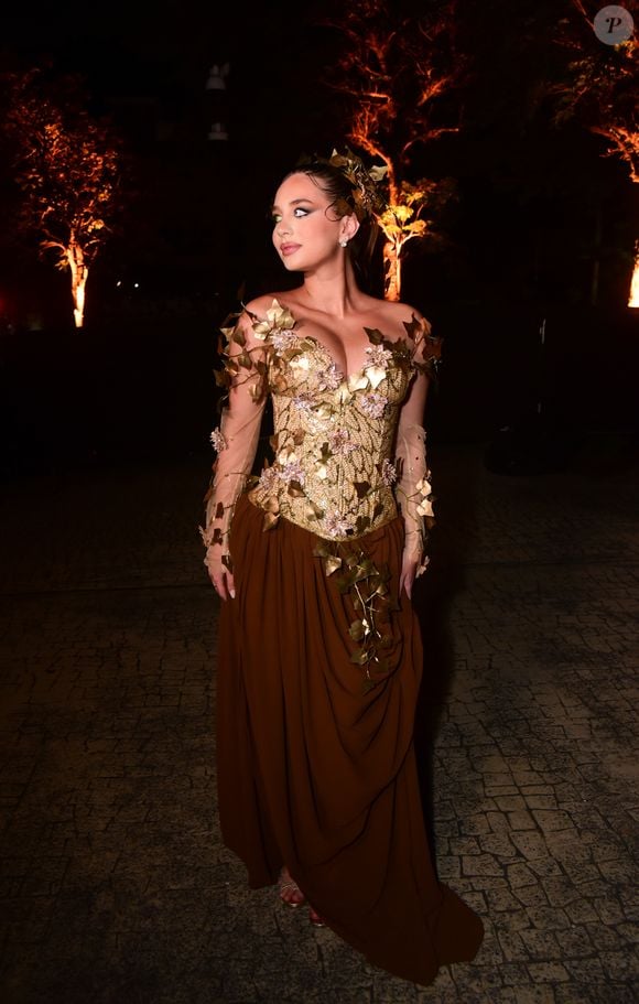Duda Rubert veste um vestido longo de gala com temática de fada e natureza. A peça possui corpete estruturado tomara que caia e mangas longas em tule transparente, decorados com bordados dourados e aplicações de folhas metálicas e pequenas flores rosa. A saia é fluida e escura, em tom marrom com caimento dramático e arranjo de folhas na cintura, harmonizando com o arranjo folheado no cabelo