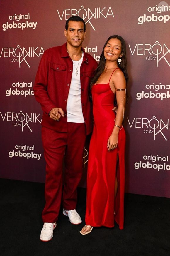 Marcelo posa ao lado de Mell Muzzillo, que usou um vestido longo vermelho-vivo de alças finas e caimento fluido. O vestido da influenciadora possui fenda lateral profunda e padronagem com suaves ondas texturizadas no tecido. Como acessórios, traz um bracelete dourado posado acima do cotovelo com pingente pendurado e sandálias rasteiras claras