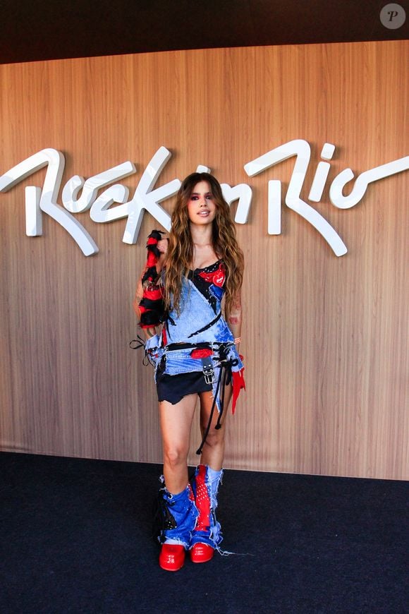 Caju ostenta um conjunto customized de inspiração country-punk em jeans destroyed e retalhos em vermelho e preto, com amarrações em ilhoses e tiras pendentes. O visual dramático traz ainda um corselet tomara que caia assimétrico, saia jeans curtíssima com barra desfiada, polainas jeans combinando sobre as botas vermelhas e bolsa transversal felpuda