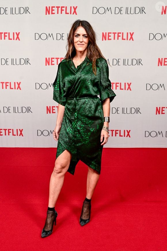 Luciana Paes veste um vestido envelope de tom verde-esmeralda com estampa animal print texturizada. A peça apresenta decote transpassado em V, mangas 3/4 e saia assimétrica com amarrações, arrematada por sapatos pretos de salto combinados com meias soquete pretas transparentes