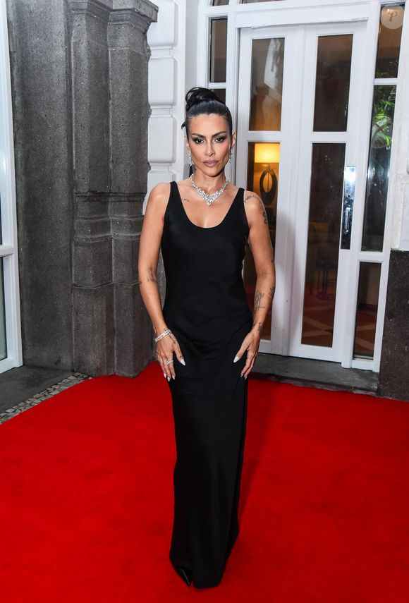 Cleo Pires apresenta uma proposta de elegância minimalista com um vestido longo de cetim preto. A peça possui decote arredondado e alças médias, com um corte enviesado que flui suavemente pelo corpo até o chão. O destaque do visual reside na joalheria de alto impacto, composta por um colar de brilhantes em formato de V, brincos coordenados e pulseiras sobrepostas