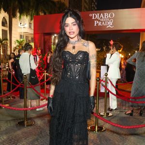 Aline Campos, ex-'BBB 26' e ex-Riscado, optou por um visual que une a ousadia da estética fetishcore com o romantismo Gótico. O look é centrado em um espartilho (corset) preto estruturado, com painéis frontais e laterais feitos de renda guipure vazada, que revela a pele de forma sofisticada e acentua a cintura. A parte inferior conta com uma saia longa de camadas de babados em tule texturizado, criando volume e movimento orgânico