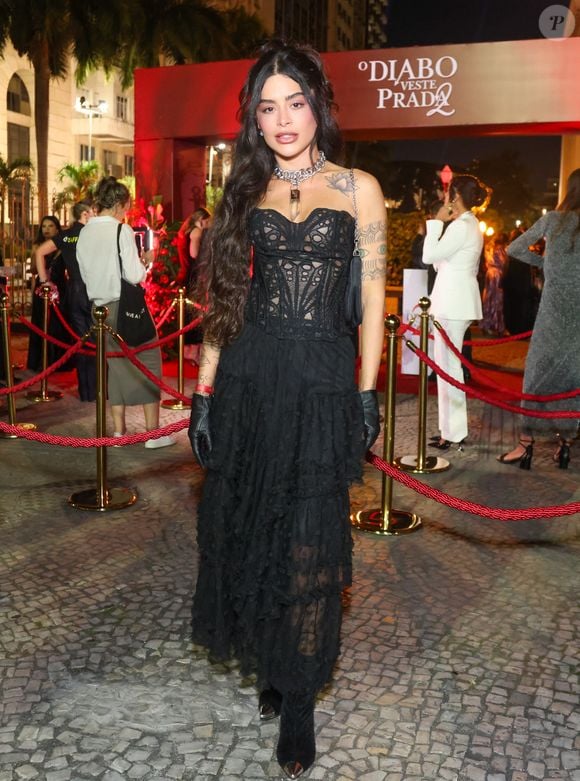 Aline Campos, ex-'BBB 26' e ex-Riscado, optou por um visual que une a ousadia da estética fetishcore com o romantismo Gótico. O look é centrado em um espartilho (corset) preto estruturado, com painéis frontais e laterais feitos de renda guipure vazada, que revela a pele de forma sofisticada e acentua a cintura. A parte inferior conta com uma saia longa de camadas de babados em tule texturizado, criando volume e movimento orgânico
