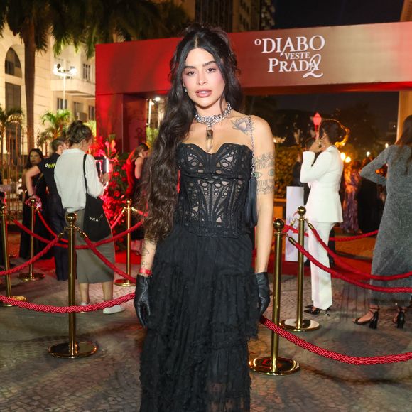 Aline Campos, ex-'BBB 26' e ex-Riscado, optou por um visual que une a ousadia da estética fetishcore com o romantismo Gótico. O look é centrado em um espartilho (corset) preto estruturado, com painéis frontais e laterais feitos de renda guipure vazada, que revela a pele de forma sofisticada e acentua a cintura. A parte inferior conta com uma saia longa de camadas de babados em tule texturizado, criando volume e movimento orgânico