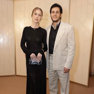 Gabriela Prioli e o marido Thiago Mansur: ela exibe um visual sutilmente audacioso com um vestido longo preto de mangas compridas em malha fina e translúcida, revelando discretamente a silhueta, arrematado por escarpins brancos de bico fino, joias minimalistas e uma bolsa de mão em couro estruturado escuro. Ele contrapõe a fluidez escura com um terno de linho texturizado em tom areia clara e modelagem oversized, combinado com uma camisa social preta aberta no peito e um lenço estampado amarrado rente ao pescoço, unindo o despojamento sofisticado à elegância formal