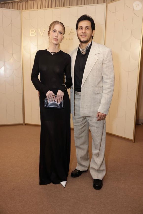 Gabriela Prioli e o marido Thiago Mansur: ela exibe um visual sutilmente audacioso com um vestido longo preto de mangas compridas em malha fina e translúcida, revelando discretamente a silhueta, arrematado por escarpins brancos de bico fino, joias minimalistas e uma bolsa de mão em couro estruturado escuro. Ele contrapõe a fluidez escura com um terno de linho texturizado em tom areia clara e modelagem oversized, combinado com uma camisa social preta aberta no peito e um lenço estampado amarrado rente ao pescoço, unindo o despojamento sofisticado à elegância formal