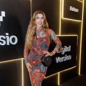 Ana Paula Minerato veste um vestido justo de comprimento mídi e mangas longas, confeccionado em tecido leve e translúcido com uma estampa botânica rica em detalhes, misturando folhagens verdes e flores em tons terrosos, avermelhados e alaranjados sobre um fundo marrom. A peça apresenta um caimento ajustado que valoriza a silhueta, complementado por um discreto drapeado na região do quadril