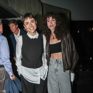 Maria Casadevall e Lua: a primeira veste alfaiataria P&B com calça ampla, camisa branca alongada sob colete curto abotoado e mocassins pretos. Ao seu lado, a acompanhante usa um cropped preto reto, jaqueta de couro marrom de corte biker, calça jeans wide leg de lavagem clara e colares com pingente