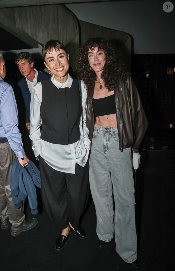 Maria Casadevall e Lua: a primeira veste alfaiataria P&B com calça ampla, camisa branca alongada sob colete curto abotoado e mocassins pretos. Ao seu lado, a acompanhante usa um cropped preto reto, jaqueta de couro marrom de corte biker, calça jeans wide leg de lavagem clara e colares com pingente