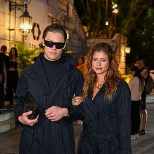 Ao seu lado, Lucas Pinheiro radicaliza no volume com uma capa oversized de nylon técnico sobre uma base de gola alta e calças pantalonas extremamente amplas, que tocam o chão. O visual de Lucas é finalizado com óculos de sol futuristas e uma clutch de couro texturizado, reforçando uma estética nômade urbana de vanguarda