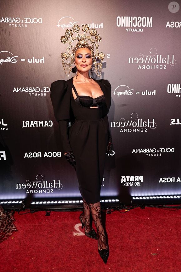 Medusa? Juliana Paes veste um vestido midi preto justo com mangas bufantes volumosas e recortes no busto, revelando um sutiã de renda preto por baixo. Ela usa luvas de ópera pretas, meia-calça de renda floral preta e sapatos de salto alto pretos de bico fino. O grande destaque é o adereço de cabeça dourado intrincado, composto por várias flores e filigranas douradas que criam um efeito de coroa imponente