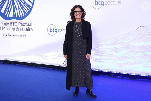 Zélia Duncan: Look moderno composto por um casaco alongado (estilo sobretudo leve) em tom verde-oliva com lapelas largas, usado sobre blusa preta de gola alta e calça social preta de modelagem reta, finalizado com sapatos pretos fechados de bico fino