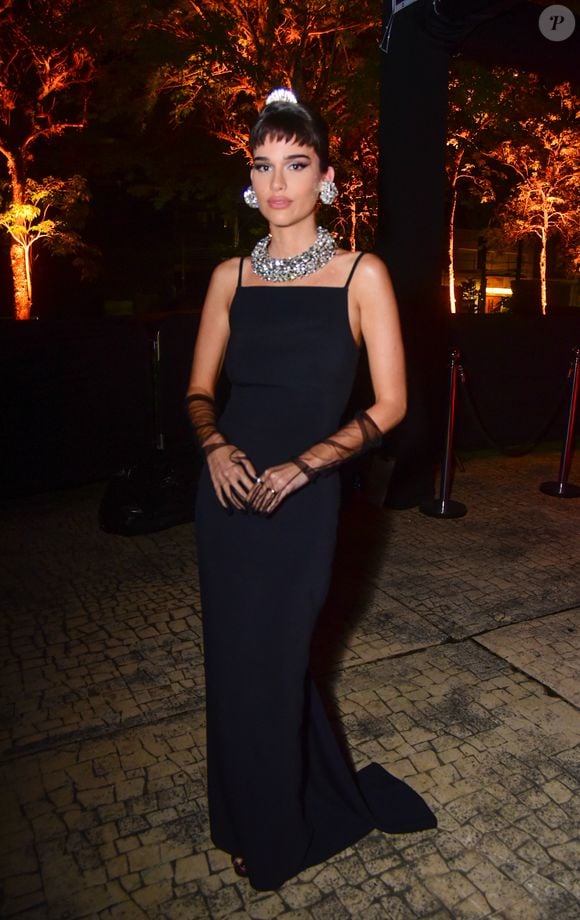 Thereza Fonseca veste um vestido longo preto liso de alças finas com decote quadrado, sem fendas aparentes. Ela acessoriza o look com luvas de ópera pretas transparentes, uma gargantilha de strass grossa, brincos grandes de strass redondos e uma pequena tiara de strass no alto do coque bagunçado. A produção evoca o estilo clássico em tom de gala