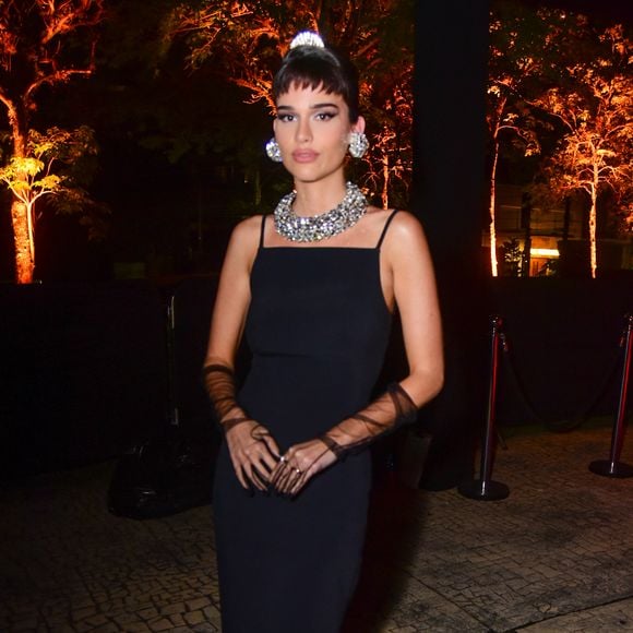 Thereza Fonseca veste um vestido longo preto liso de alças finas com decote quadrado, sem fendas aparentes. Ela acessoriza o look com luvas de ópera pretas transparentes, uma gargantilha de strass grossa, brincos grandes de strass redondos e uma pequena tiara de strass no alto do coque bagunçado. A produção evoca o estilo clássico em tom de gala. Mariane Noronha fez o cabelo da atriz com produtos Sebastian Professionals.