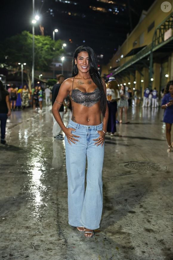 Pocah escolheu um top frente-única em malha de brilho/cristais prateados com decote degagê, combinado a uma calça jeans de cintura baixa e modelagem reta em lavagem clara. O visual traz detalhes como braceletes finos no braço, brincos de argola grandes, salto alto prateado aparente e cabelos pretos soltos alisados