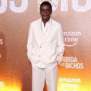 Seu Jorge aposta em um terno monocromático off-white de caimento relaxado, composto por blazer transpassado com bolsos utilitários e lapelas amplas, combinado a uma camisa acetinada levemente aberta. A calça de alfaiataria reta complementa o conjunto fluido, arrematado de forma contemporânea por tênis brancos casuais